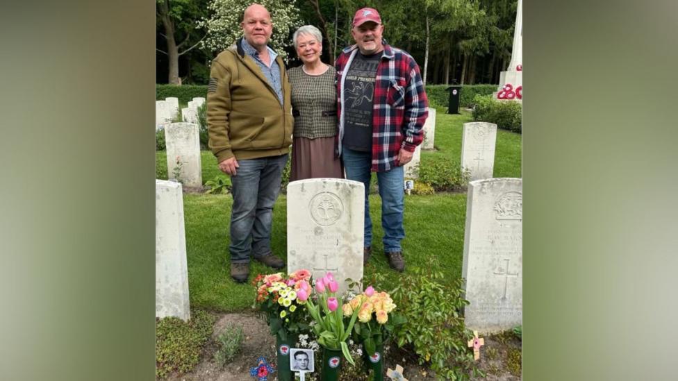 Margaret is standing in between Bart (left) and Bernie (right) in the cemetery. Bart is wearing a blue shirt, jeans, and a khaki green zip-up jacket. Margaret is wearing a brown skirt and green jacket. Bernie is wearing a baseball hat, blue jeans, a grey t-shirt and red, navy and blue check print jacket. There are flowers on Maynard Powell's grave.
