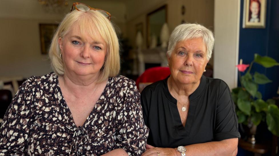 Julie (left) has medium-length blonde hair, glasses on her head, and she is wearing a brown printed top. Her mother, Margaret (right) has short white hair and is wearing a black t-shirt. They are both smiling.
