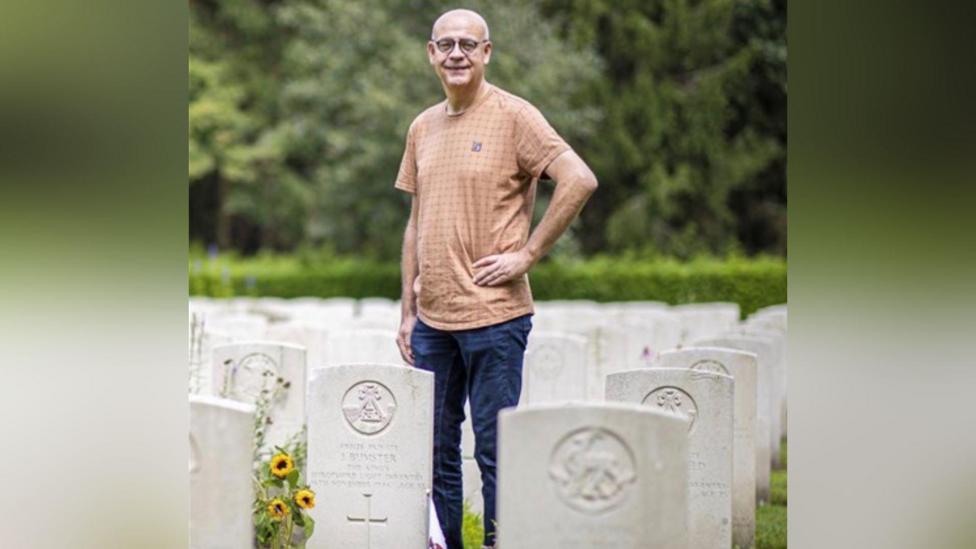 Theo Vervoort standing in the cemetery in Venray. He is wearing a light orange t-shirt and navy blue jeans. He is wearing glasses. He is smiling and has his hand on his hip.