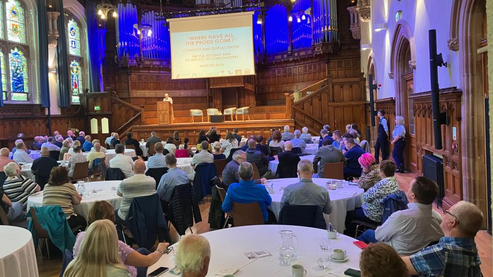 A number of people are sat around circular tables in Derry's Giuildhall. A person is pictured at the podium.