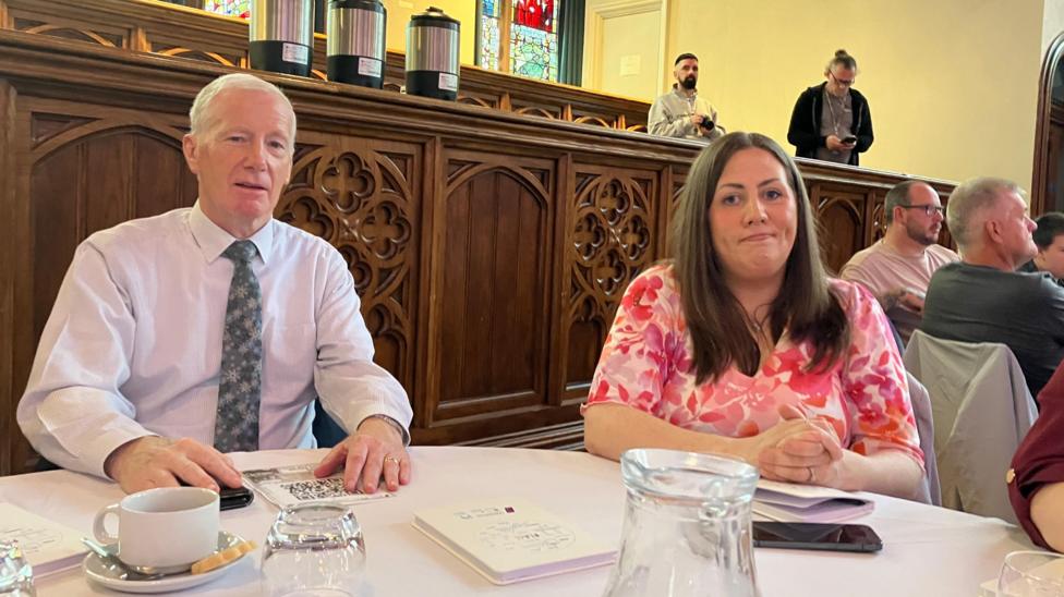 DUP MP Gregory Campbell and DUP MLA Julie Middleton are sat at a circular table with a white tablecloth. Gregory Campbell is wearing a white shirt and colourful green tie. Julie Middleton is wearing a pink and white dress.