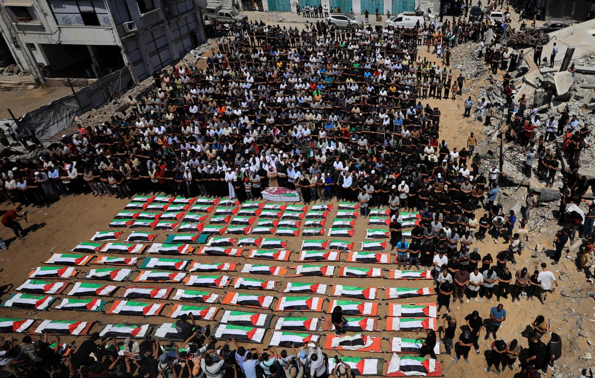 Flags draped over the bodies of dozens of Palestinians as a crowd of mourners pray nearby.