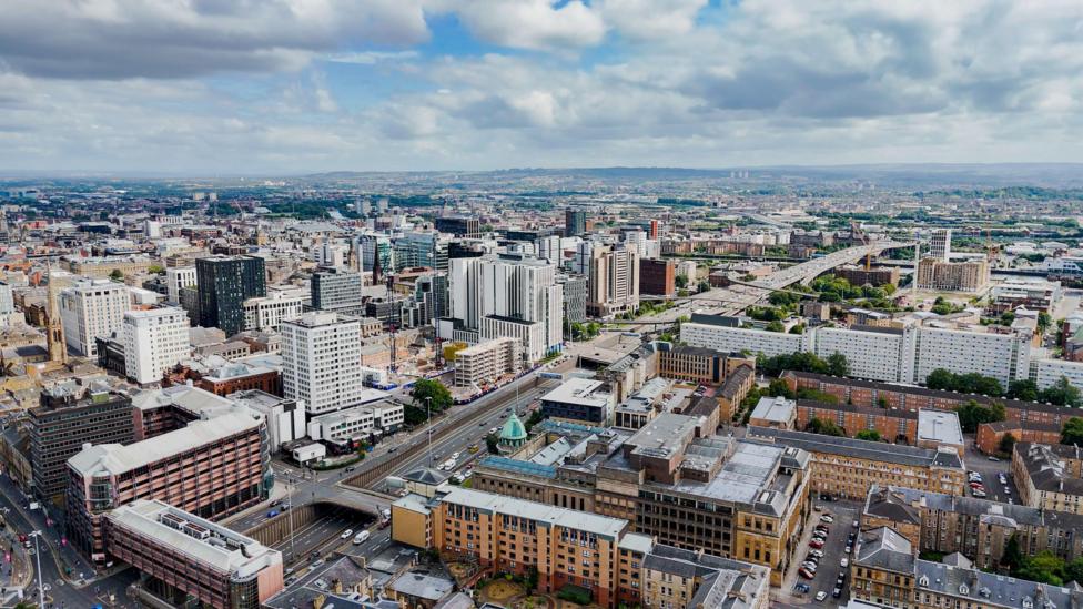 An aerial overhead view of the city of Glasgow. The city’s motorway cuts through the urban core with busy traffic and surrounding modern and historic architecture. There are a mixture of high-rise offices, traditional sandstone buildings and civic landmarks with domed roofs. 
