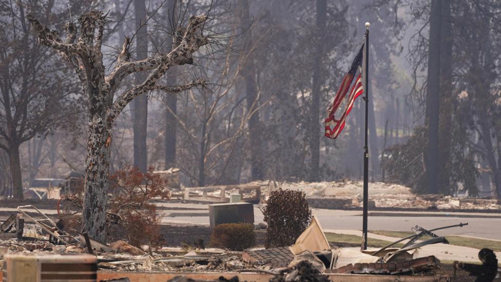 A burned United States flag hangs from a charred flagpole - burned trees and remnants of destroyed properties can be seen on either sides of the road and the air is hazy