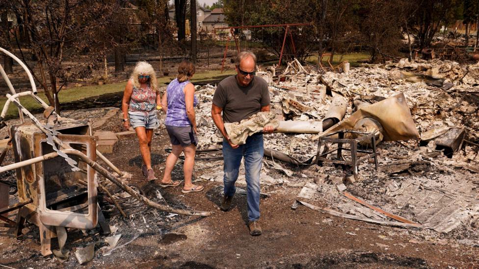 A man carrying a small statue as he walked through burned-out rubble and ashes, with a child's swing seen in a garden in the background - two women wearing vests and shorts are behind him, one turning to look at the ruins