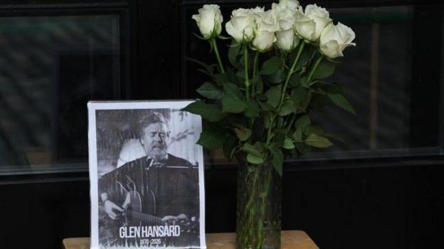 A black and white picture of Glen Hansard sits on a table with a vase of white roses beside it