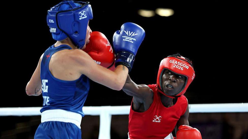 Angel Katushabe, wearing all red boxing gear, including headgear and gloves, throws a punch at her opponent, wearing all blue, during a boxing match.