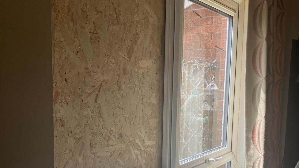 A piece of wood is placed over a smashed window. to the right of the wood a smashed window pane can be seen.