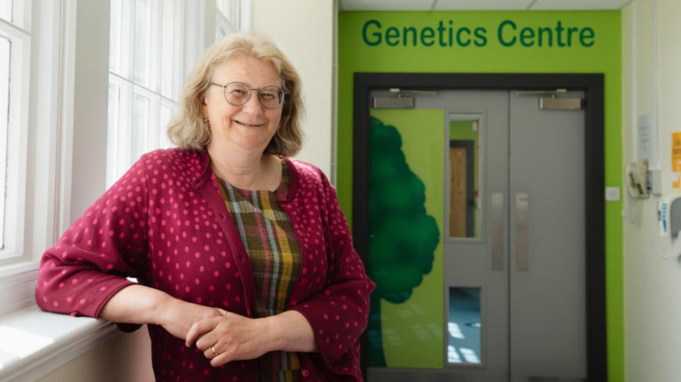Prof Zosia Miedzybrodzka is standing in a brightly lit corridor, in front of a green door with the words 'Genetics Centre' above. She has light brown hair and is wearing glasses, a tartan top and a red cardigan. She is smiling at the camera