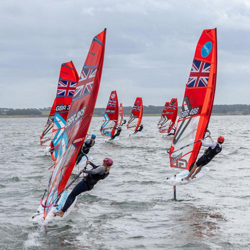 A series of wind surfers with bright red sails carrying the Union Jack