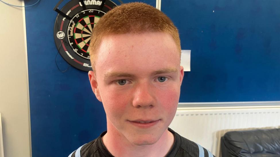 Shane O'Neill has cropped red hair and is wearing a sports top. A dart board on the wall behind him.