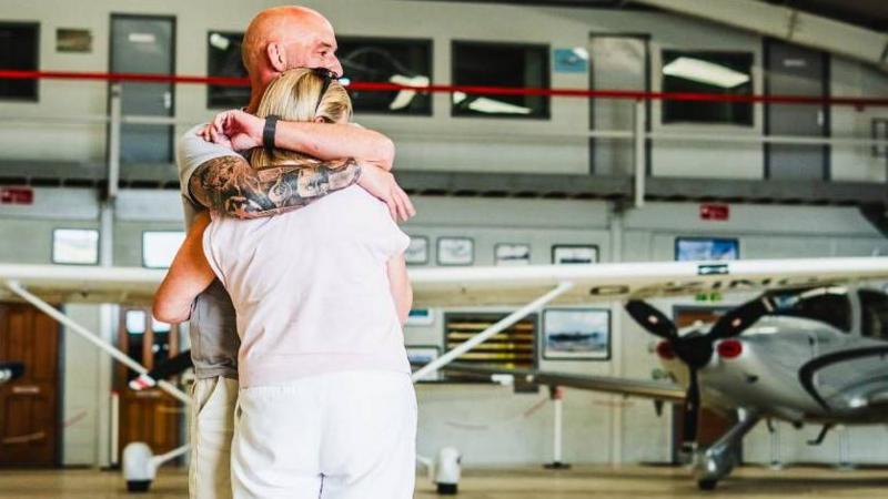A man and woman are seen hugging. There is a plane in the background, on the right of the photo.