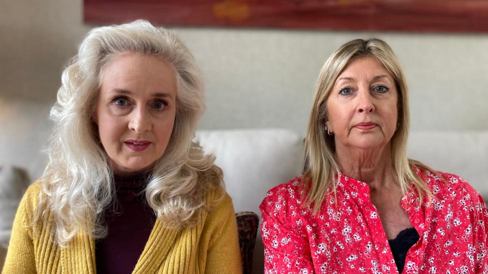 Two women are sat beside each other on a white sofa. Nicola Bannon, a woman with shoulder-length wavy white hair, is sitting on the left wearing a mustard cardigan over a black top. Lynne Darcy has shoulder-length straight blonde hair and is wearing a pink patterned collarless shirt over a black top.