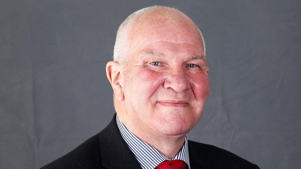 A bald-headed man with short, cropped grey hair smiles into the camera. He is in a dark suit with a striped shirt and a red tie. The background is grey.