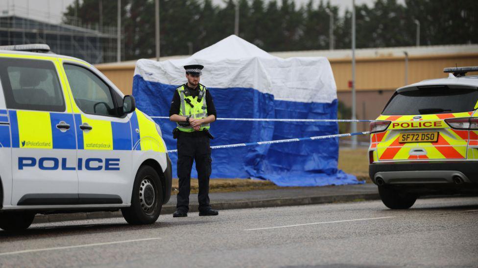 A police officer standing in front of a police cordon and blue forensic tent and two police vehicles in Arbroath.