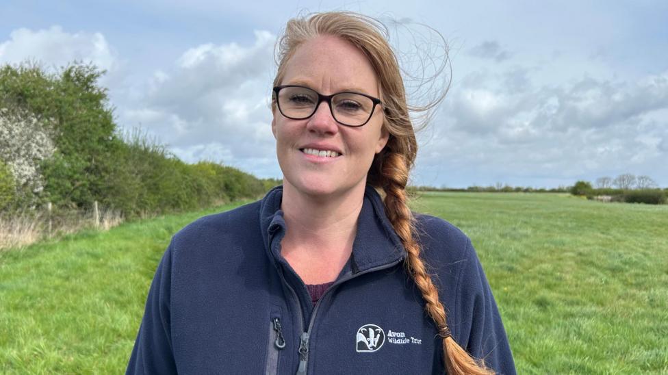 Shelly Easton wearing a navy blue zip fleece with Avon Wildlife Trust on the front and the Trust's badger logo. She is standing near a hedgerow in a field.