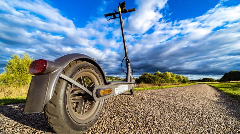 A close-up of an e-scooter on a sunny day. The vehicle is black with two black wheels, a narrow black steering column, a red rear light and an amber side light on the wheel. A gravel path lined by grass and trees stretches into the distance in front of the e-scooter.