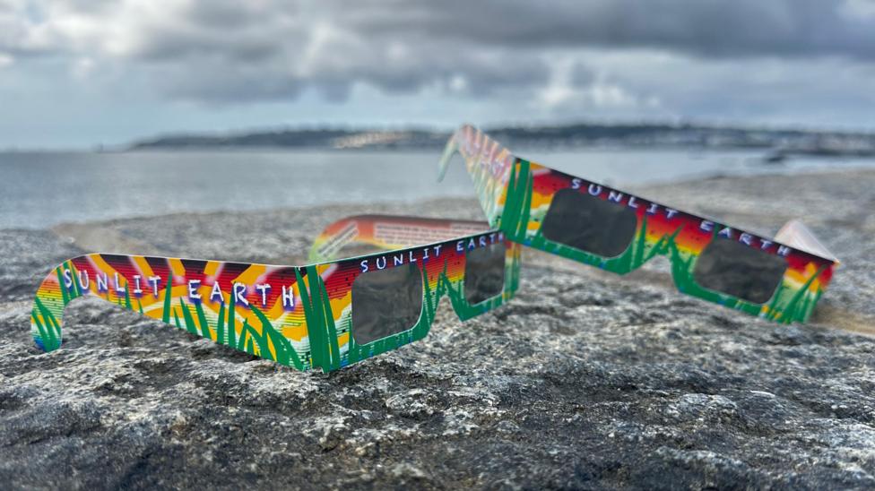 Two pairs of brightly coloured solar eclipse carboard glasses. One pair is resting on the other on top of a granite wall. A blurred view of Guernsey is in the background.