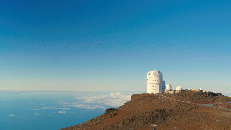 The image shows an observatory - a white, dome-like structure, perched on a peak overlooking the ocean. Scattered clouds are visible below the observatory and the sky above is clear and blue. This is the NSF Inouye Solar Telescope - the most powerful solar telescope in the world.