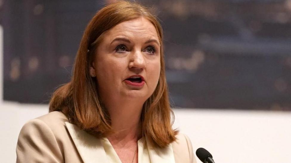 A woman with red shoulder length hair talks into a microphone. She is wearing a cream and white jacket.