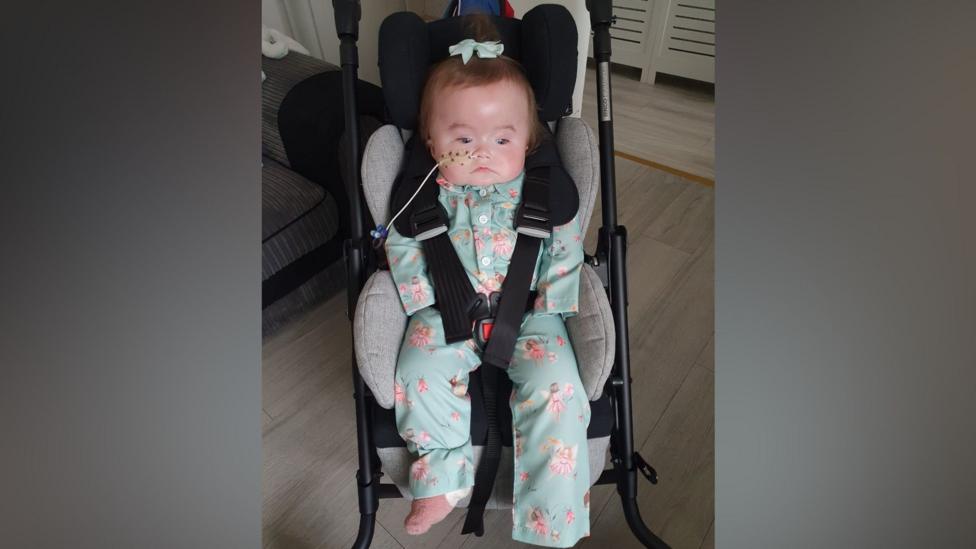 Lila is pictured in her buggy - which is a dark colour with grey padding for her to sit on. She is strapped into it. She's wearing a matching green floral top and trousers and there's a bow on top of her dark hair. A tube is in her right nostril. The buggy is in a room with a grey wooden floor.