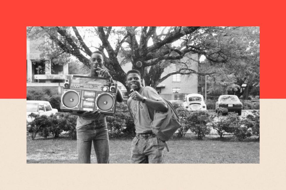 Two young men stand by a boombox and smile. The photograph is in black and white