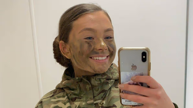 A mirror selfie of Jaysley Beck wearing a camoflauge jacket with green and brown face paint on her face. Her brown hair is tied up in a slick back low bun and she is smiling at the camera.