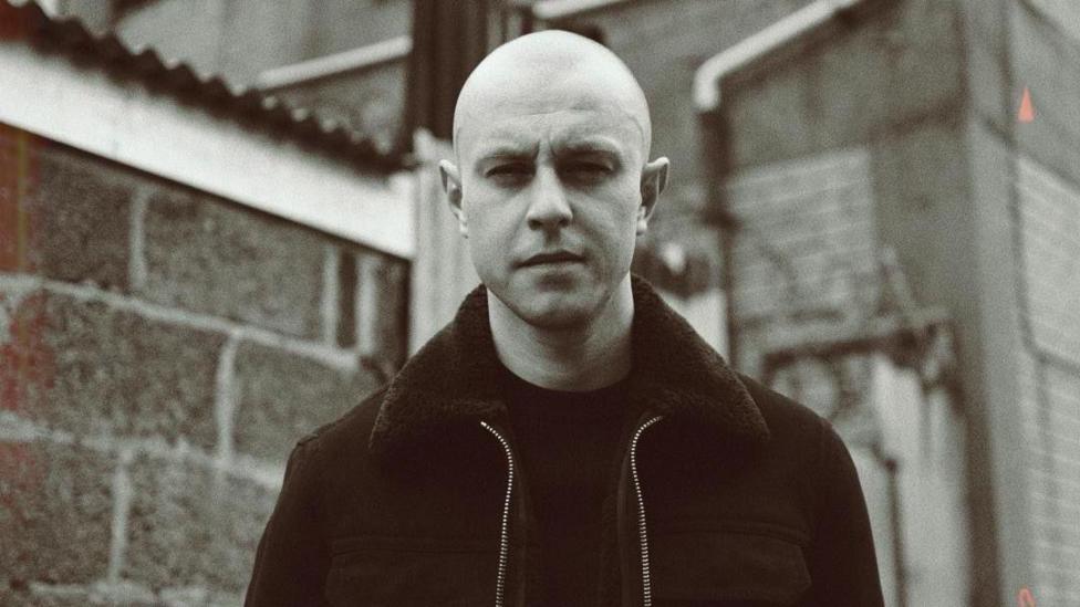 A black and white picture of Young Spencer. He has a shaved head and is wearing a dark jacketand top 