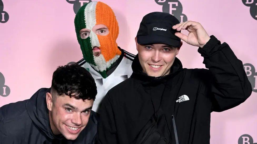 The free members of kneecap two of who are dressed in dark clothing and the one in the middle is wearing a white sports top and Irish tricolour balaclava
