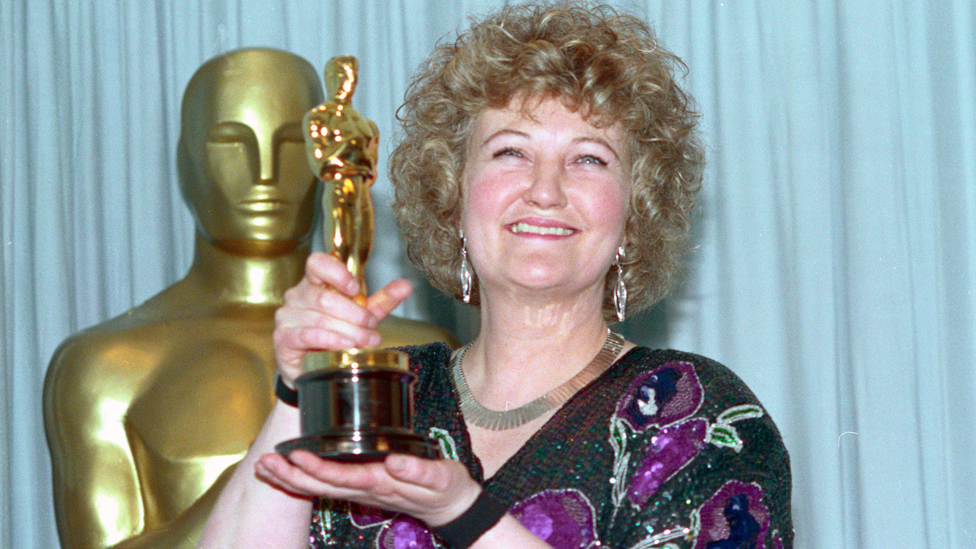 Brenda Fricker hold a small golden statue. It has a black base which is held by her left hand, her right grips the statues legs. She wears a black purple sequenced top. She has medium length blonde hair and smile to the camera, wearing dangling silver earings.