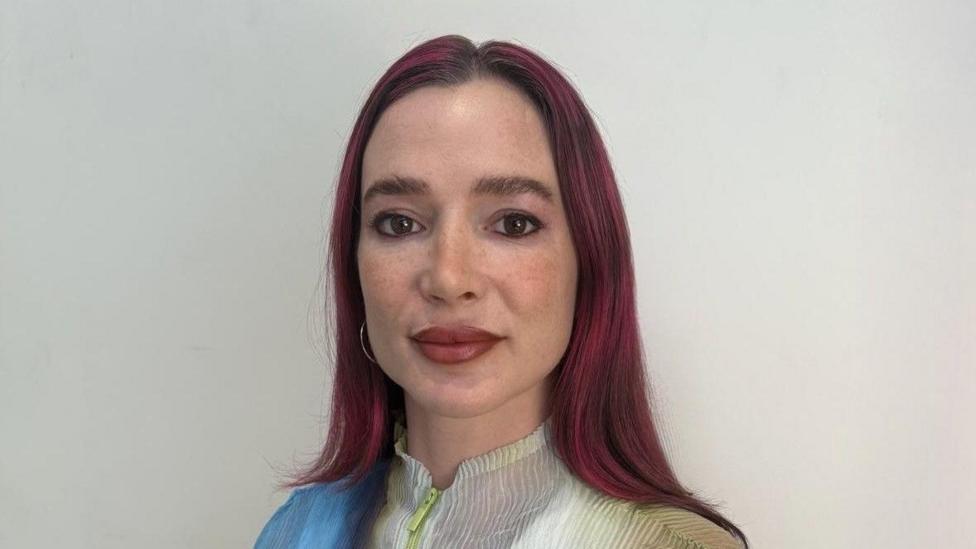 A woman with long dark red hair looks at the camera