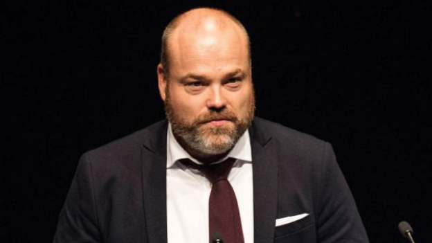 a bald man with a brown beard wearing a dark suit, white shirt and purple tie
