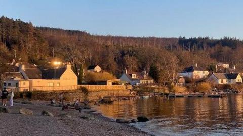 The Dores Inn and nearby homes are on a shore of Loch Ness. The properties are bathed in sunlight.