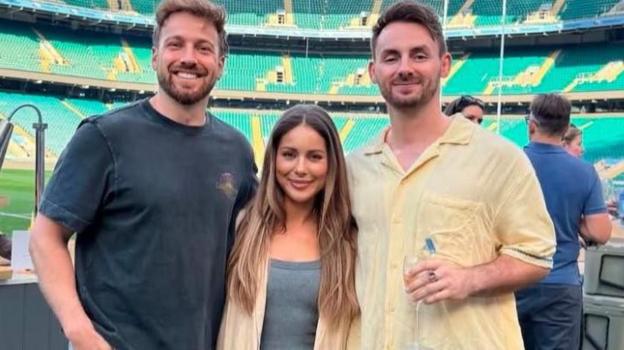 Sam and Louise Thompson along with Scott are stood at an event in a stadium. Behind people are chatting and food is being served while the group smile at the camera.