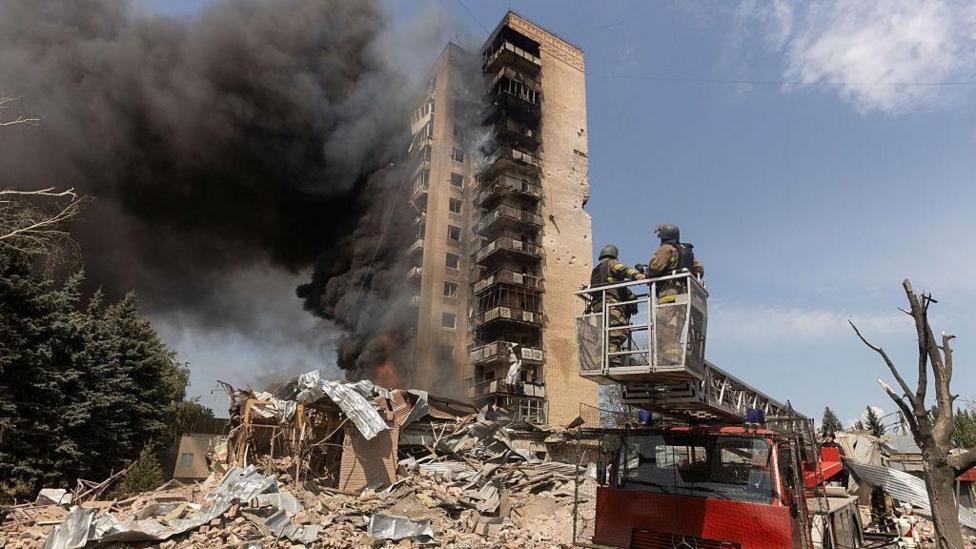 A half-ruined building is burning. It is surrounded by brick rubble as black smoke billows out the side of it. A red truck has two firefighters dressed in brown overalls on top of it observing. The sky is clear and blue. A green tree sits to the left of the picture.
