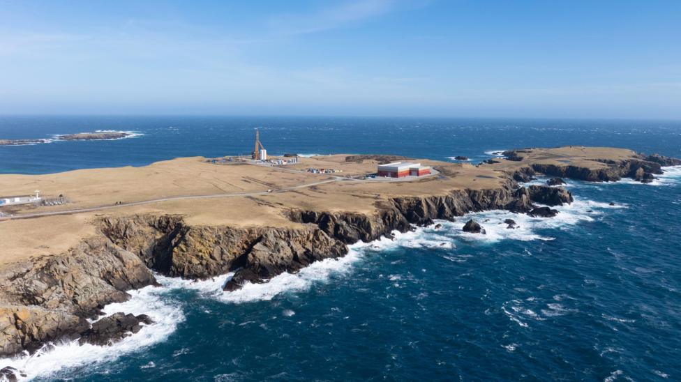 An aerial view of a long flat peninsula surrounded by dark blue sea. The coastline is rocky and the sky is blue. The island is brown and sparsely populated by several industrial buildings and a tall launch pad.