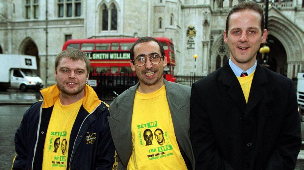 Three men stand in front of the Royal Courts of Justice in London with a red London bus also behind the,. They are smiliang wearing yellow tshirts