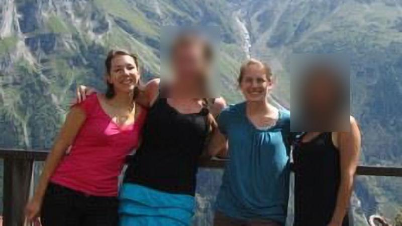 A group of four people stand together on a viewing platform with a railing, posing in front of a dramatic mountain landscape featuring steep rocky cliffs and a waterfall. Two faces are blurred. One person wears a bright pink top, another a black top with a blue skirt, another a blue top, and another a dark outfit. The scene is outdoors in daylight with rugged green and gray mountains in the background.