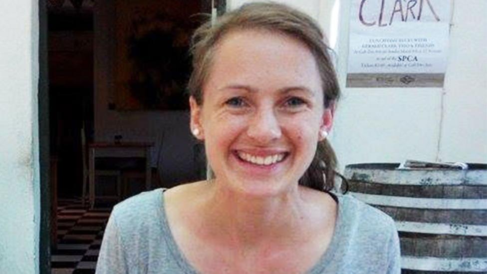 A woman with long brown hair, tied back in a ponytail, smiling at the camera. She is wearing pearl-like earrings and a scoop neck grey top.