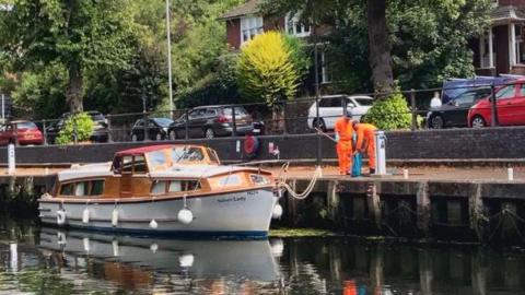 Anglian Water workers wearing orange high-vis protective clothing use a net to collect fish and put them in a blue bag, while stood along the bank of a river next to a motor cruiser