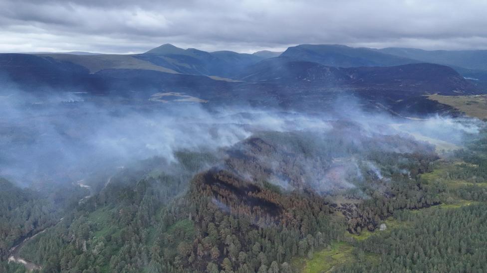 Smoke rises from a large area of forestry covering low hills. Higher hills and mountains are in the background.