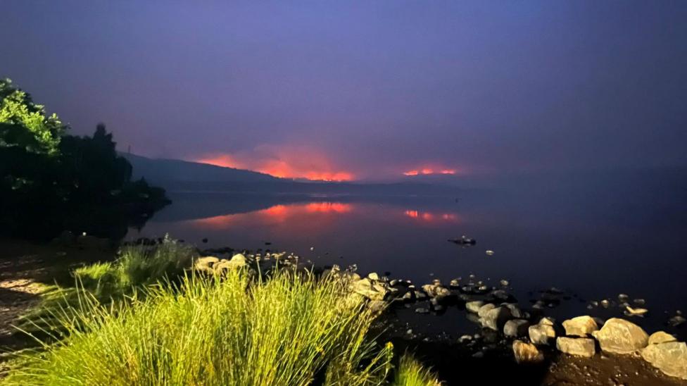 It is dark and flames glow orange on the horizon. The flames are reflected in the mirror-like surface of Loch Morlich.