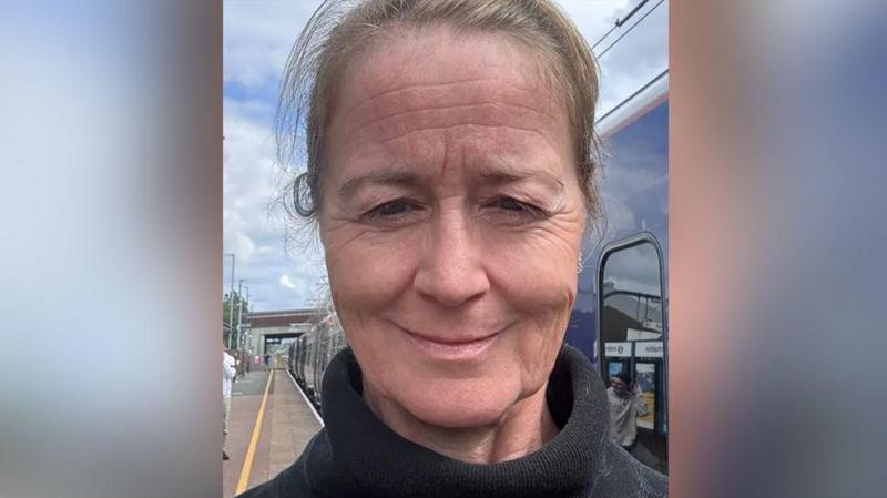 Sarah Nelson selfie on a railway station platform after having to come off a train. She has light brown hair worn back in a ponytail and wears a black polo neck jumper. She is smiling.