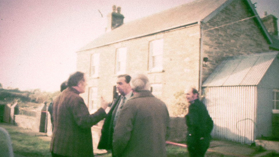 A still from archive footage of police officers outside Ffynon Samson farmhouse in December 1976. DCS Pat Molloy is stood in the centre of the group, wearing a suit and talking to his colleagues, as a uniformed officer looks on.