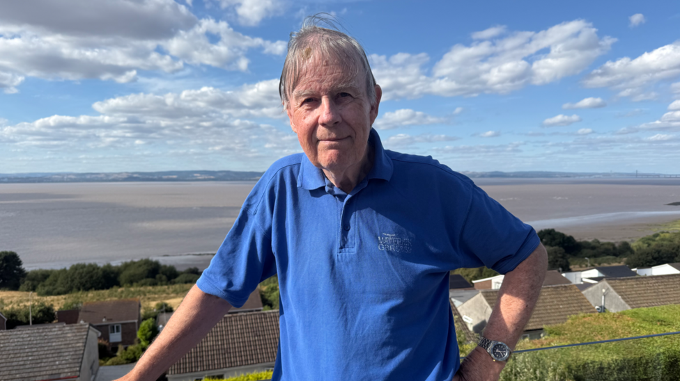 Huw looks at the camera, with the sea behind him. He has short grey hair and wears a blue polo shirt.