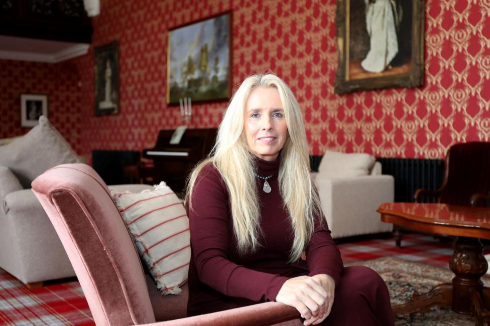 Suzanne has long blonde hair and is wearing a burgundy top. She is sitting in an armchair in a large room with ornately decorated red wallpaper.