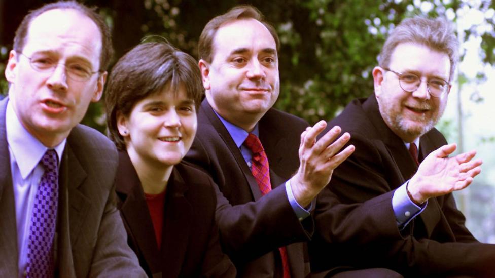 John Swinney and Nicola Sturgeon stand in a row with Alex Salmond and Michael Russell. They are all in business wear and are smiling and waving.