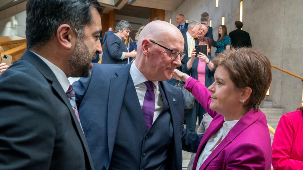In the grey and light wood lobby of the Scottish Parliament, Humza Yousaf, John Swinney and Nicola Sturgeon are standing talking. Swinney is warmly embracing Sturgeon, who is staring beyond him. Yousaf stands off to the side. There is a crowd of people on the stairs behind them. Someone is taking a picture.