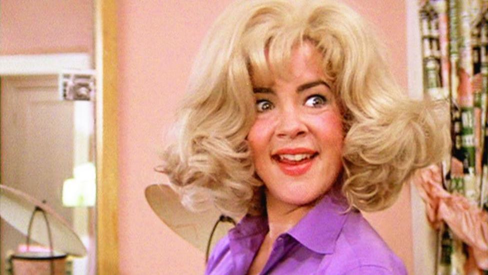 A smiling woman in a blonde wig wearing a purple shirt