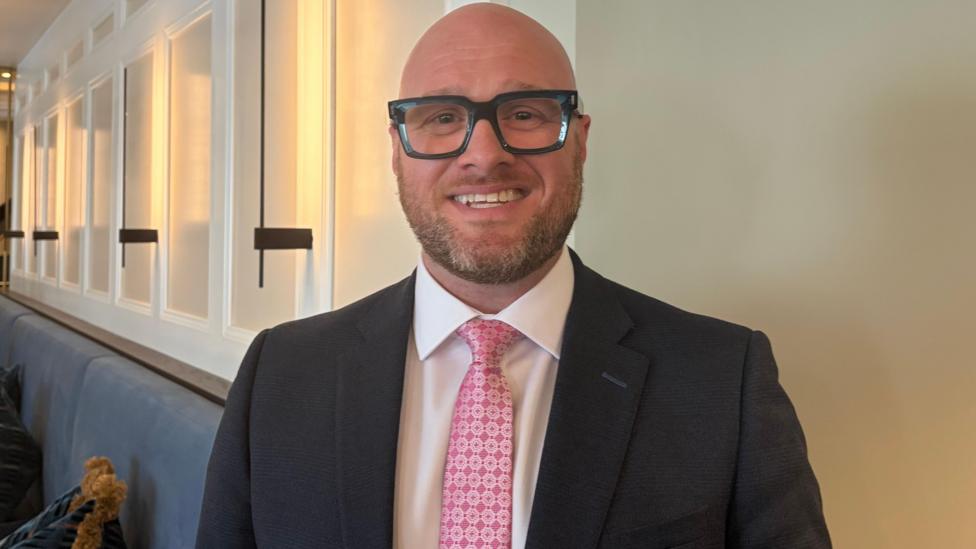 A man in a black suit with white shirt and pink tie. He is bald and wearing black glasses.